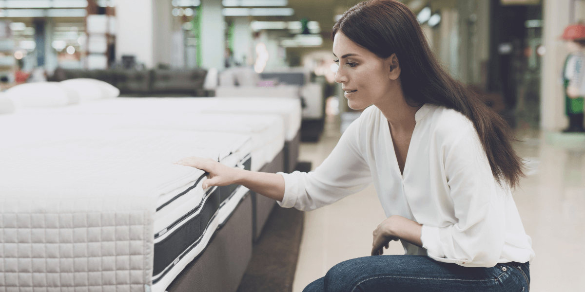 A woman kneels beside a mattress, examining its surface and features closely.